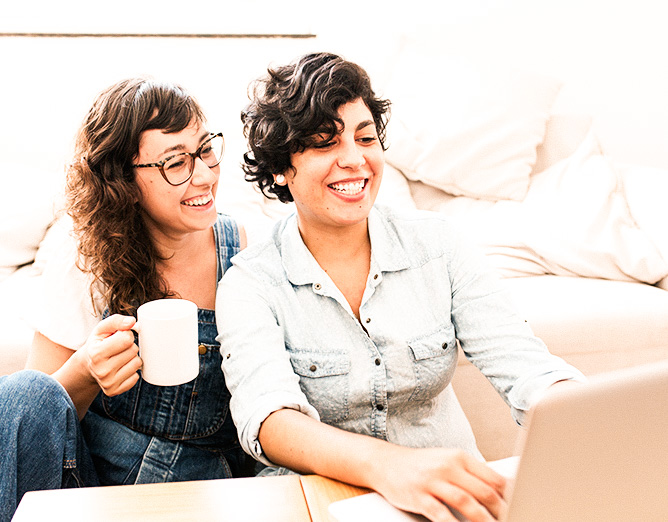 Image of two smiling women sitting on a couch and looking at a laptop screen.