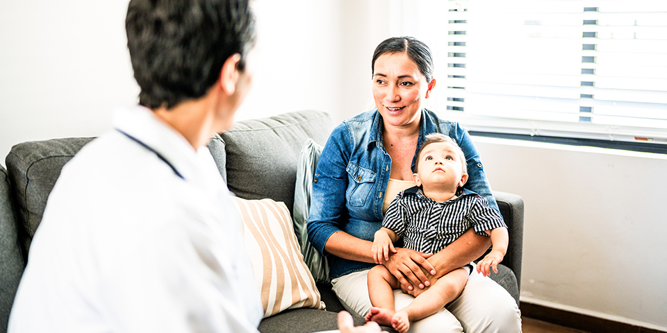 A mother holding her baby on her lap sit on a couch talking to a medical professional.